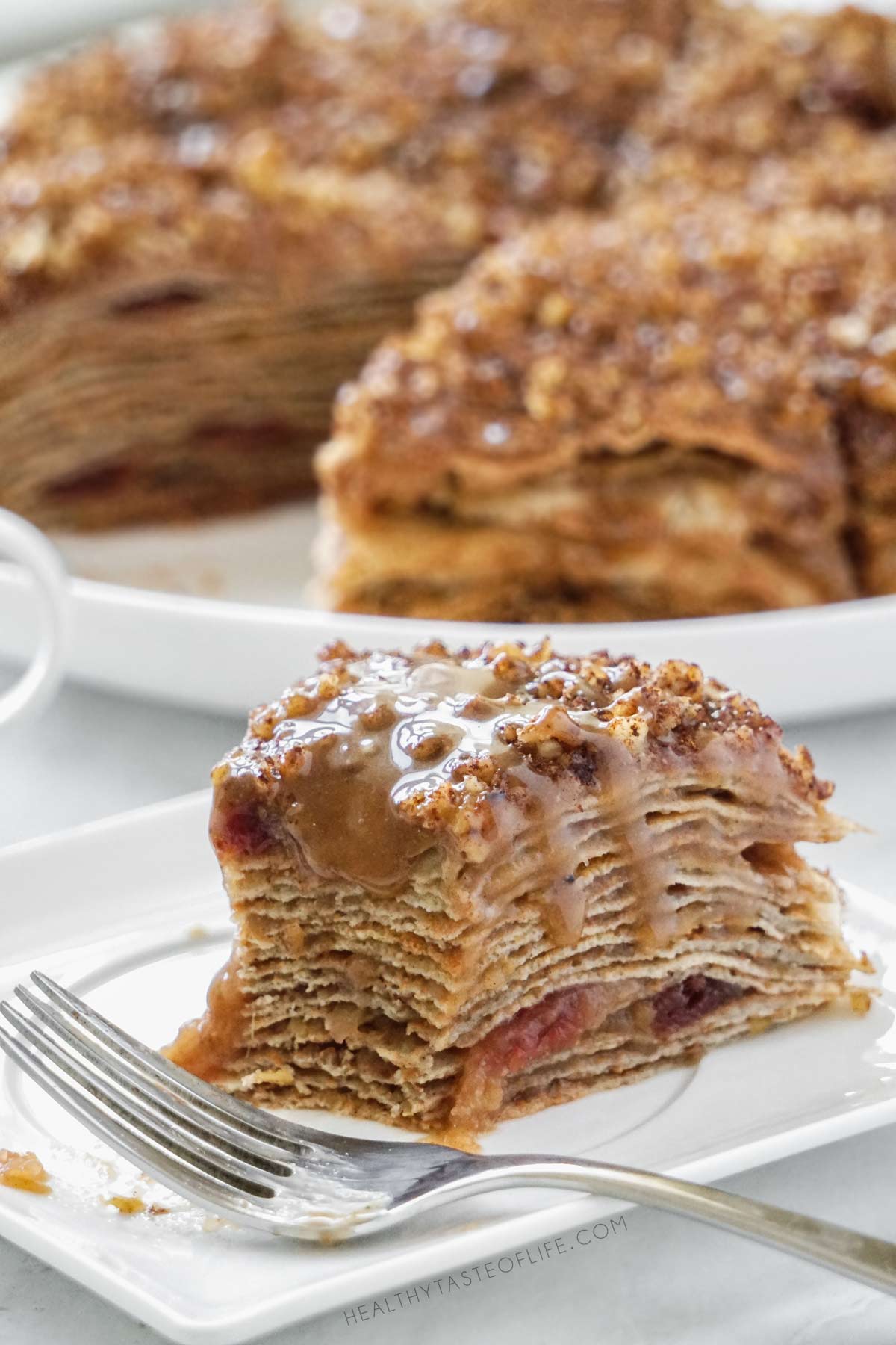 A a portion of apple crepe cake separated from the main slice showing the inside texture of gluten free crepes, cooked apple filling and dairy free caramel sauce dripping
